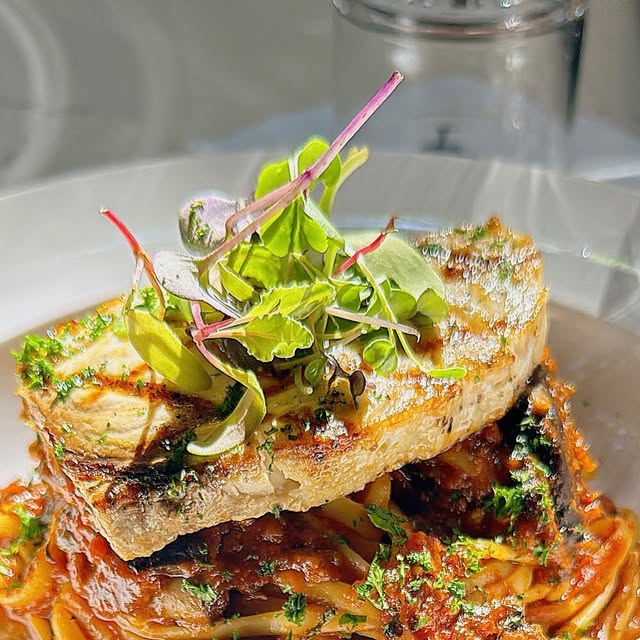 Grilled swordfish over linguine puttanesca on a white plate, crowned with microgreens